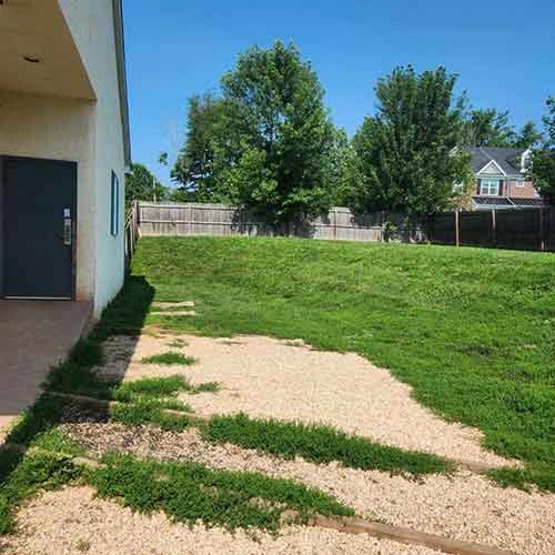 A grassy yard sloping gently upwards, bordered by trees and a wooden fence. In the foreground, a path of gravel leads from a building with a door. The sky is clear and sunny, indicating bright weather.